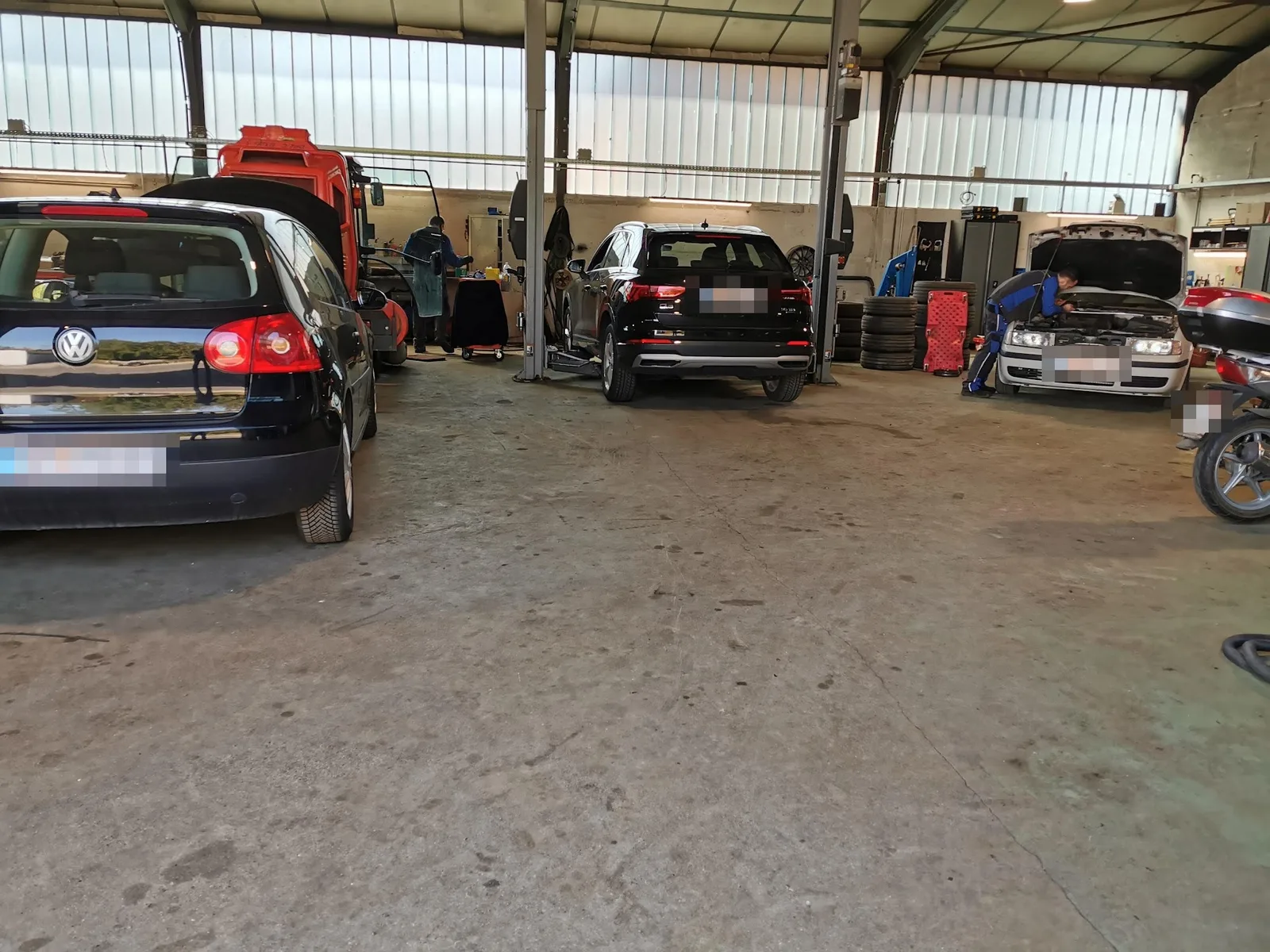 Werkstatthalle mit mehreren Arbeitsplätzen, ein Mitarbeiter arbeitet am offenen Motorraum eines Pkw
